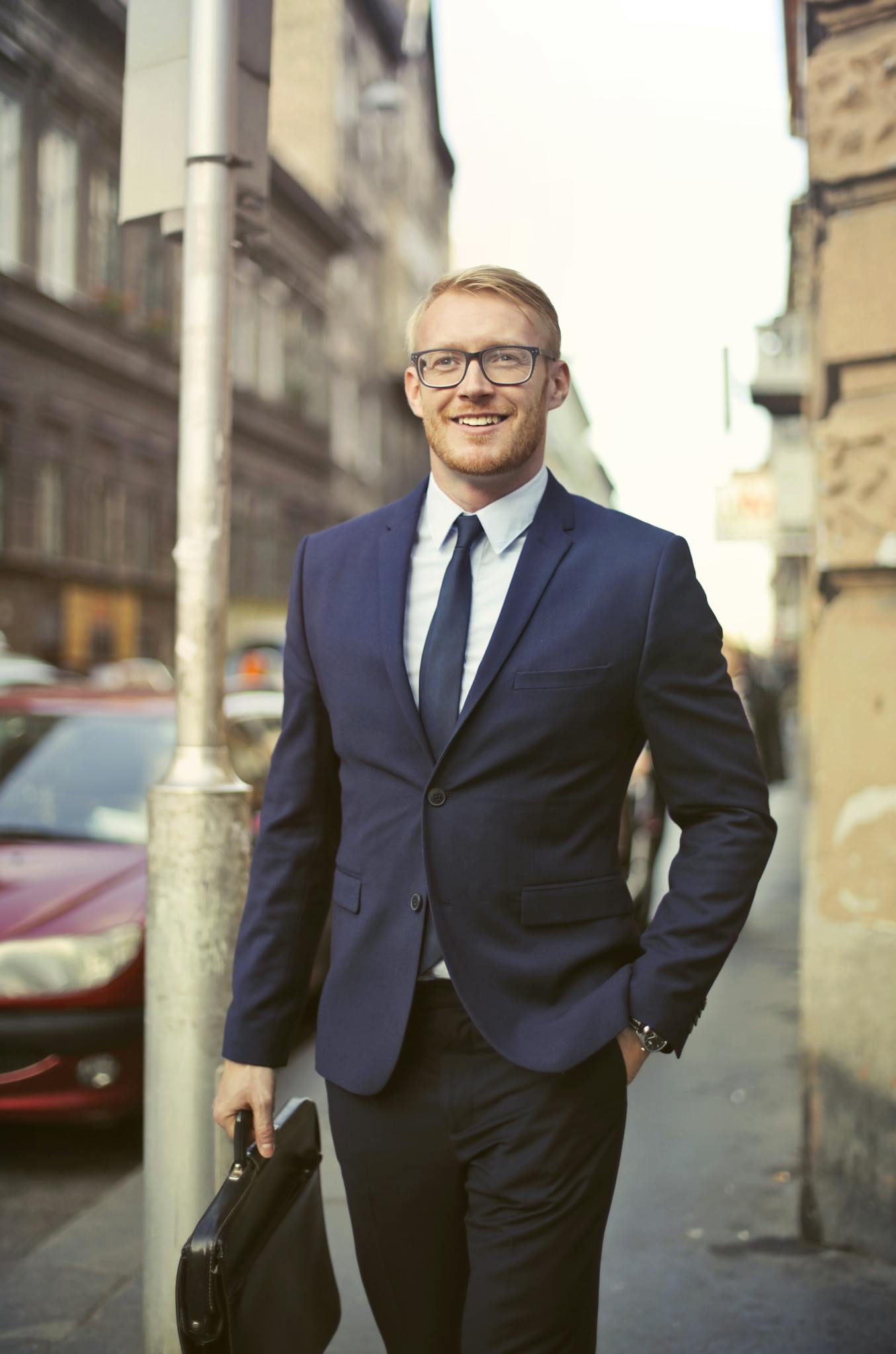 Smiling businessman in suit with briefcase walking on urban street with parked cars.